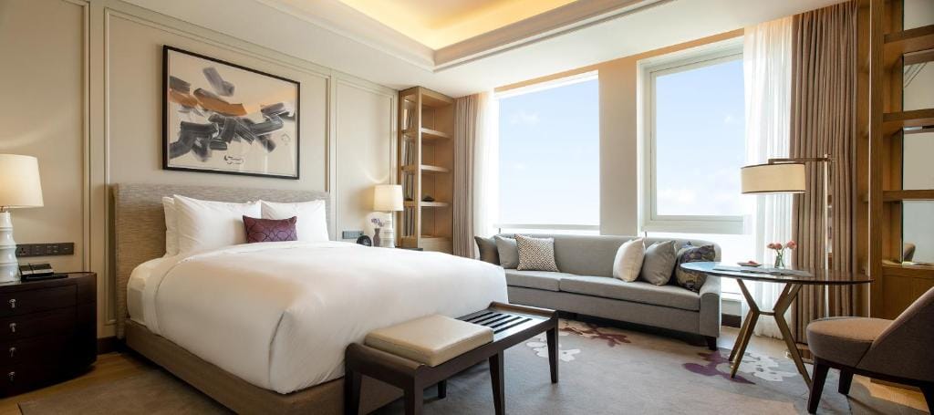A modern hotel room with a large bed, a bench at the foot, a sofa by the window, a round table with a lamp, and wall art above the bed. Natural light streams in through large windows with sheer curtains.