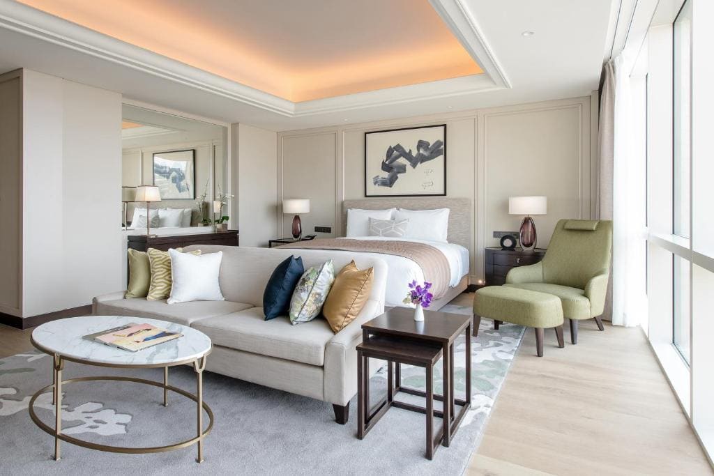 A modern hotel room with a large bed, a beige sofa with colorful pillows, a round marble coffee table, a green armchair with ottoman, two bedside tables with lamps, a large window, and soft lighting.