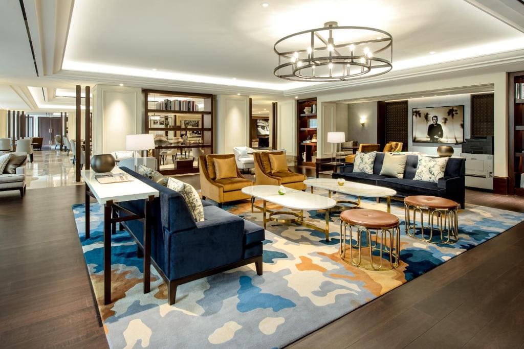 A modern, elegant lounge with velvet sofas, armchairs, marble coffee tables, decorative stools, and a patterned rug. Bookshelves and artwork decorate the space, and soft lighting creates a warm, inviting atmosphere.