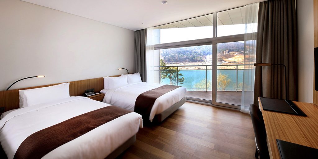 Modern hotel room with two double beds, brown accents, wooden floor, large window and balcony overlooking a lake and wooded hills in the background. A desk and TV are on the right side of the room.