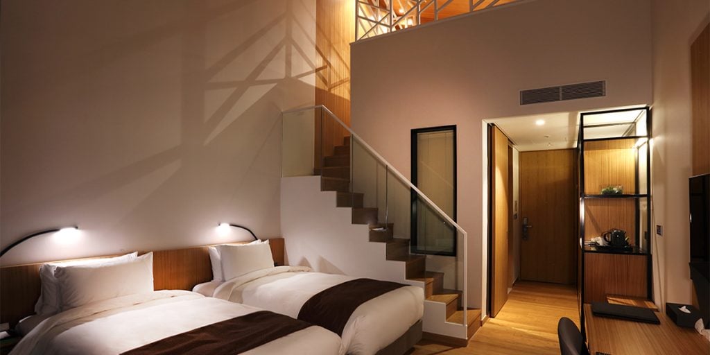 Modern hotel room with two twin beds, brown blankets, wall-mounted reading lights, a loft area accessible by stairs with a glass railing, and a hallway with wood flooring and a black mini-fridge and kettle.