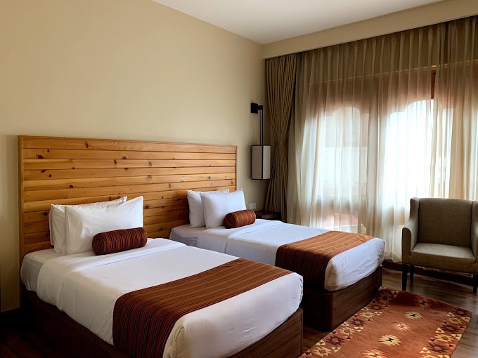 A hotel room with two single beds, each with white bedding, brown striped runners, and cylindrical cushions, wooden headboard, a window with sheer curtains, an armchair, and a patterned rug on a wooden floor.