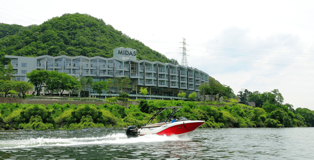 A red and white speedboat travels on a river in front of a modern building labeled MIDAS, set against a green, tree-covered hillside under a partly cloudy sky.