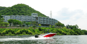 A red and white speedboat travels on a river in front of a modern building labeled MIDAS, set against a green, tree-covered hillside under a partly cloudy sky.