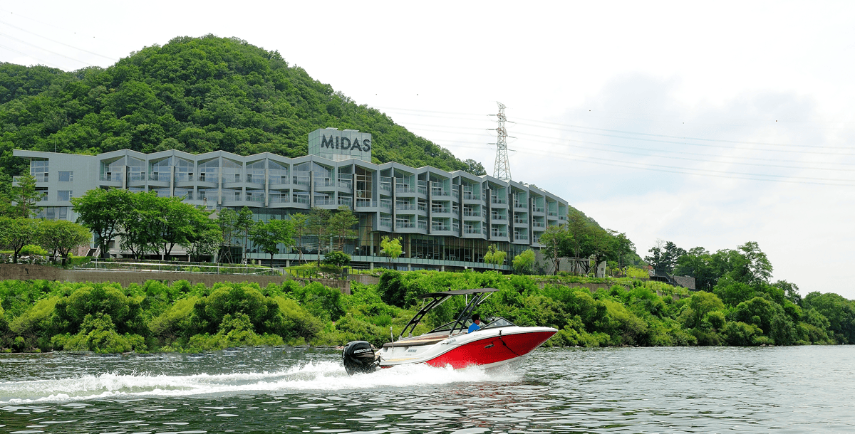 A red and white speedboat travels on a river in front of a modern building labeled MIDAS, set against a green, tree-covered hillside under a partly cloudy sky.