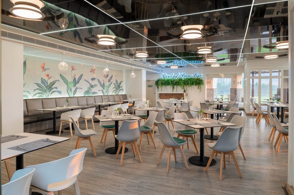Modern restaurant interior with light wood floors, gray and white chairs, tables set for dining, a botanical mural on the wall, and a neon “find a paradise” sign above greenery in the back.