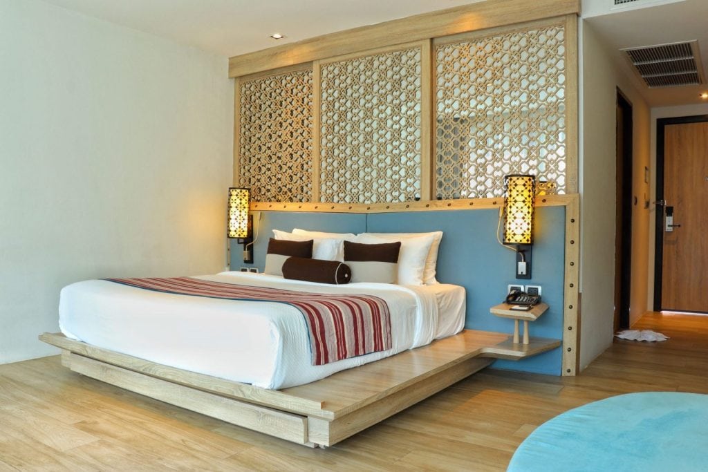 A modern hotel room with a low wooden bed, white bedding with striped accents, decorative pillows, ornate partition behind the headboard, bedside lamps, light wood floor, and a door in the background.
