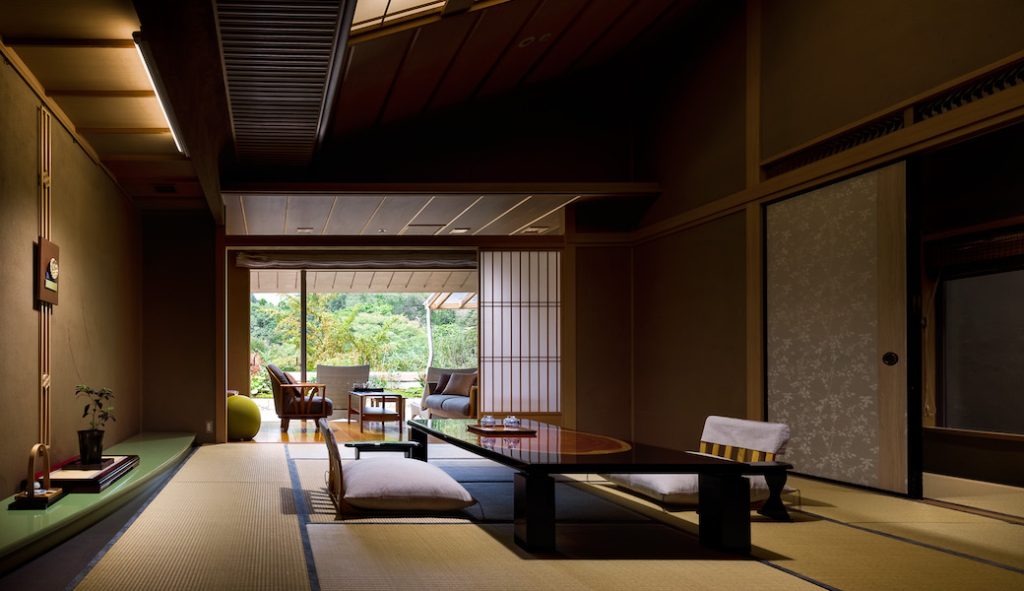 A traditional Japanese tatami room with a low wooden table, floor cushions, sliding shoji doors, and an open view to a sunny terrace with greenery outside. The room features minimalistic decor and warm lighting.