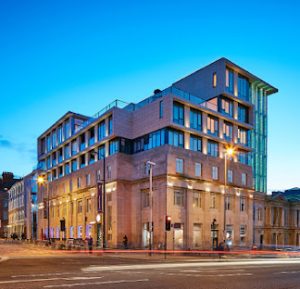 Modern multi-story building with large windows and angular architecture at a street corner, illuminated by streetlights at dusk, with a clear blue sky in the background. Light trails from passing vehicles are visible on the road.