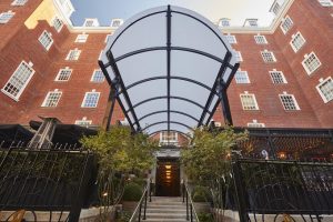 Entrance to a red-brick building with tall windows, black metal gate, steps leading up, and a large transparent canopy overhead, surrounded by greenery.