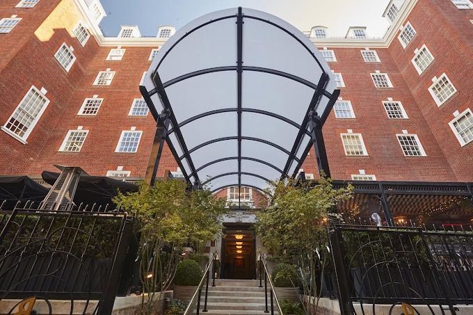 Entrance to a red-brick building with tall windows, black metal gate, steps leading up, and a large transparent canopy overhead, surrounded by greenery.