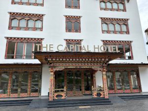 The entrance of The Capital Hotel features traditional Bhutanese architectural designs and intricate colorful patterns, with large windows and a sign displaying the hotel’s name above the doorway.