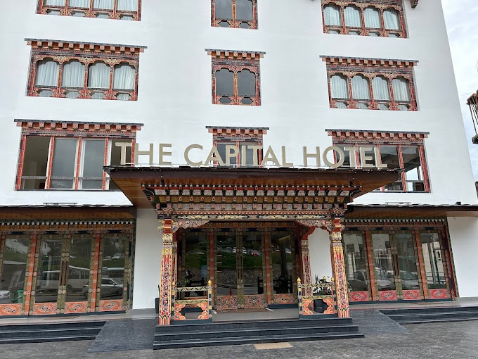 The entrance of The Capital Hotel features traditional Bhutanese architectural designs and intricate colorful patterns, with large windows and a sign displaying the hotel’s name above the doorway.