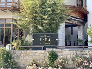 A hotel entrance with a sign reading The Pema by Realm in gold letters, surrounded by stone walls, plants, flowers, and a modern building with wooden and glass features in the background.