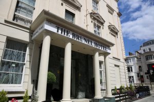 The entrance of The Brunel Hotel, featuring tall columns, potted plants, and large windows on a cream-colored building, with a clear blue sky and nearby buildings in the background.