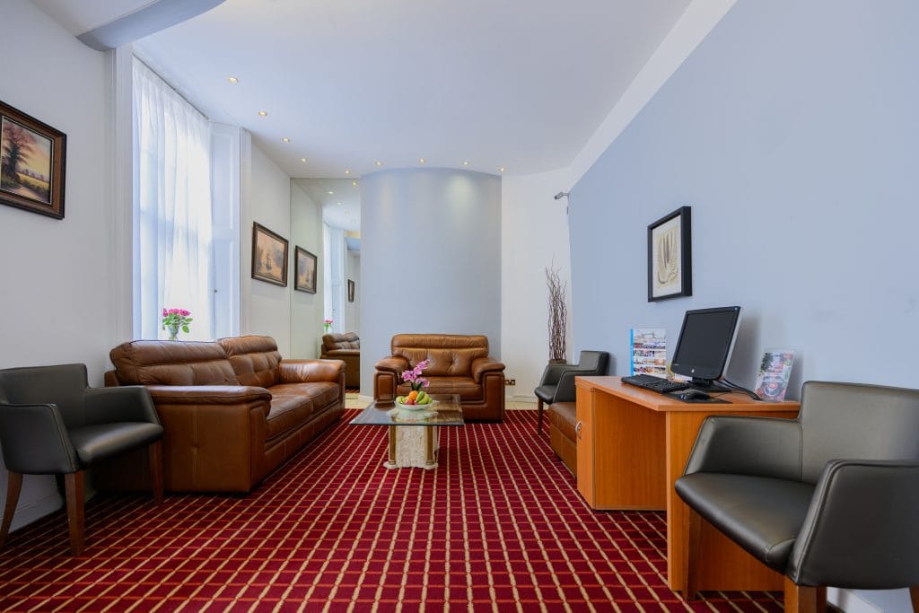 A modern waiting room with brown leather sofas, black chairs, a red patterned carpet, a computer on a wooden desk, potted flowers, and paintings on light blue walls.