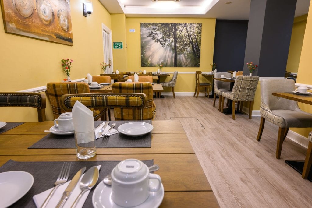 A cozy, modern dining room with wooden tables set for breakfast, upholstered chairs, yellow walls, and artwork including a large nature photo. The room is softly lit and has a relaxed, inviting atmosphere.