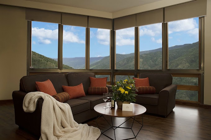 A cozy living room with a dark brown sofa, orange and brown cushions, a cream blanket, a round coffee table with a plant, and large windows displaying scenic mountain views under a blue sky with clouds.