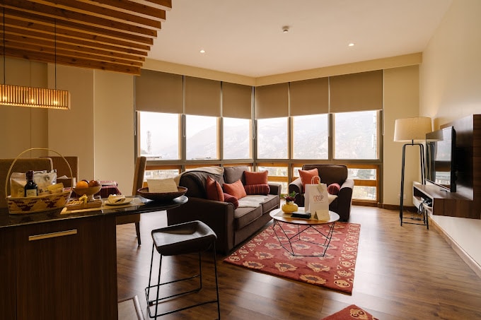 Modern living room with large windows, a gray sofa, armchair, red patterned rug, and a TV mounted on the wall. Kitchen counter with stools and a breakfast basket in the foreground. Warm lighting and wooden floor.