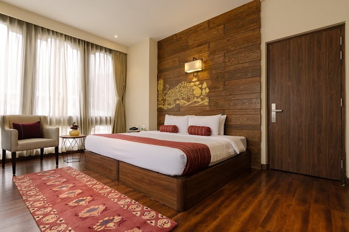 A modern hotel room with a large bed featuring white linens and red accents, wooden floors, a matching wood-paneled wall, a wall lamp, a cozy chair, side table, and large windows with sheer curtains.