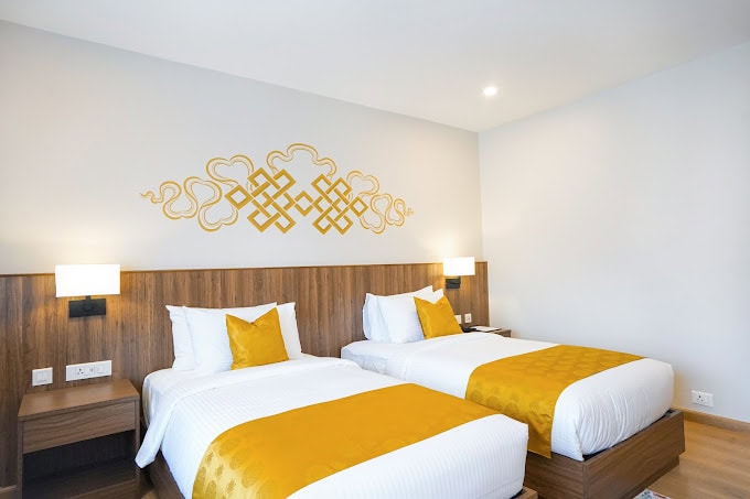 A modern hotel room with two single beds, each with white linens and golden yellow runners and pillows. The headboard is wooden, and above it is a decorative golden wall pattern. The room has light walls and wooden flooring.