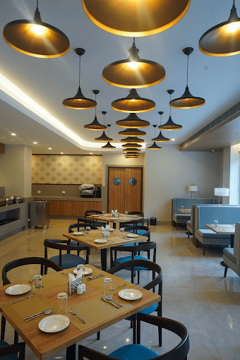Modern restaurant interior with wooden tables set for dining, black chairs, blue booth seating along the wall, and multiple black and gold pendant lights hanging from the ceiling. The space is bright and neatly arranged.