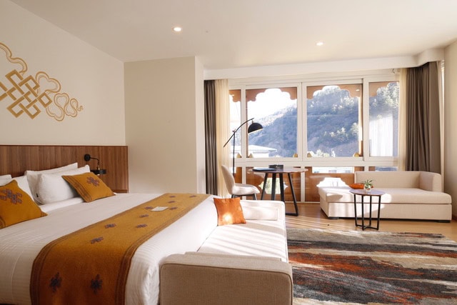 A bright hotel room with a large bed, cushions, a cozy sofa, a desk with a lamp by the window, and a decorative rug. Sunlight streams through wide windows, offering a view of hills outside.