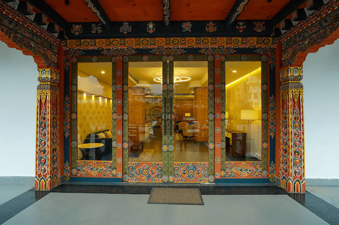 Ornately decorated wooden doorway with colorful traditional patterns, leading to a modern, warmly lit interior with glass doors and visible seating inside.