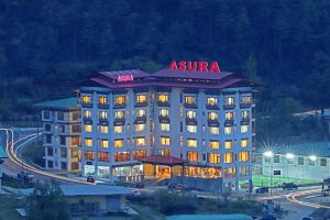 A brightly lit multi-story hotel named ASURA sits amid a hilly, forested area at dusk, with cars and light trails visible on surrounding roads.