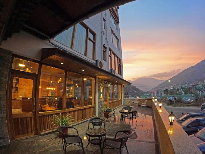 Outdoor seating area of a café or restaurant at sunset, with wicker tables and chairs, warm indoor lighting, mountain views, and a colorful sky in the background.