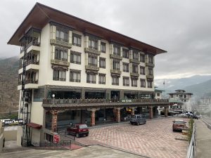 A four-story hotel with traditional Bhutanese architectural details, parked cars in front, and mountains visible in the background under a cloudy sky.
