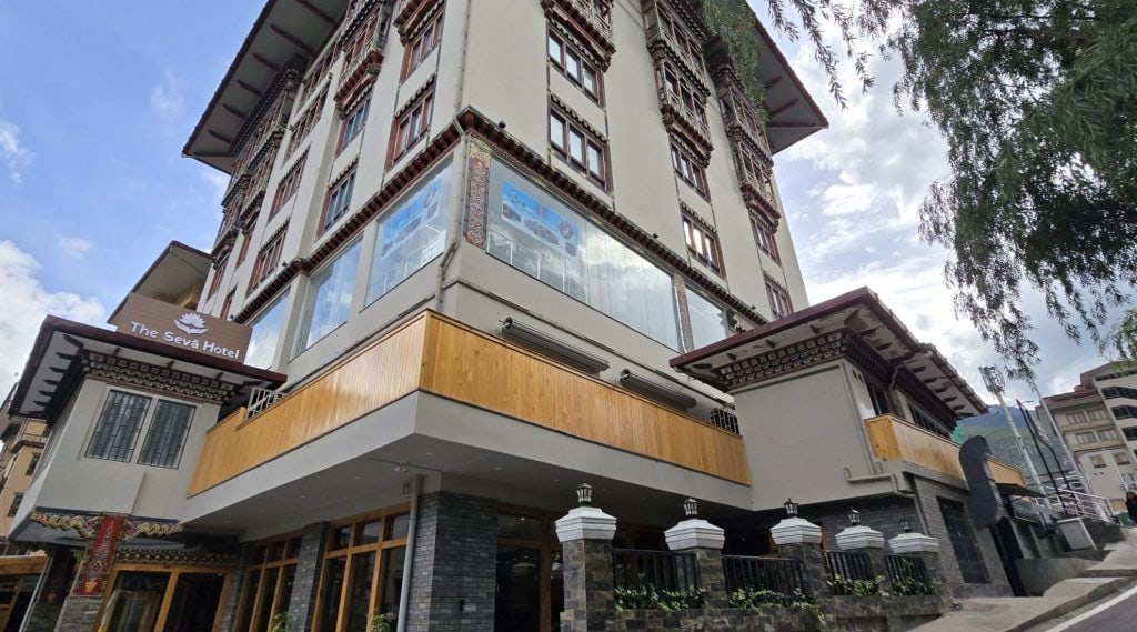 A multi-story hotel with traditional Bhutanese architectural features, including ornate window frames and painted designs, viewed from a low angle against a partly cloudy sky.