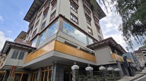 A multi-story hotel with traditional Bhutanese architectural features, including ornate window frames and painted designs, viewed from a low angle against a partly cloudy sky.
