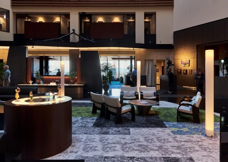 A modern hotel lobby with contemporary furniture, soft lighting, art displays, and a patterned carpet. People are visible in the background, and large windows let in natural light.