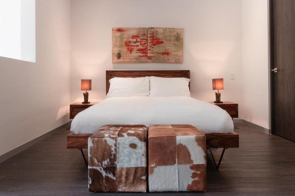 A modern bedroom with a wooden bed frame, white bedding, flanked by two nightstands with lamps, abstract art above the bed, and two cowhide ottomans at the foot of the bed.