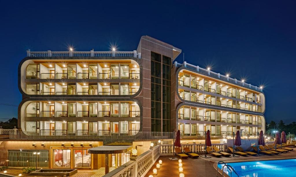 A modern, multi-story hotel building with balconies, illuminated at night. The foreground features a swimming pool surrounded by lounge chairs and umbrellas.