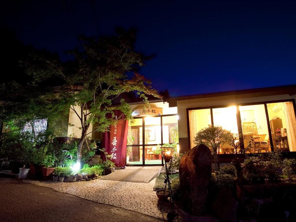 A cozy building warmly lit from within at night, featuring large windows, a red curtain at the entrance, and lush greenery surrounding the entrance path.