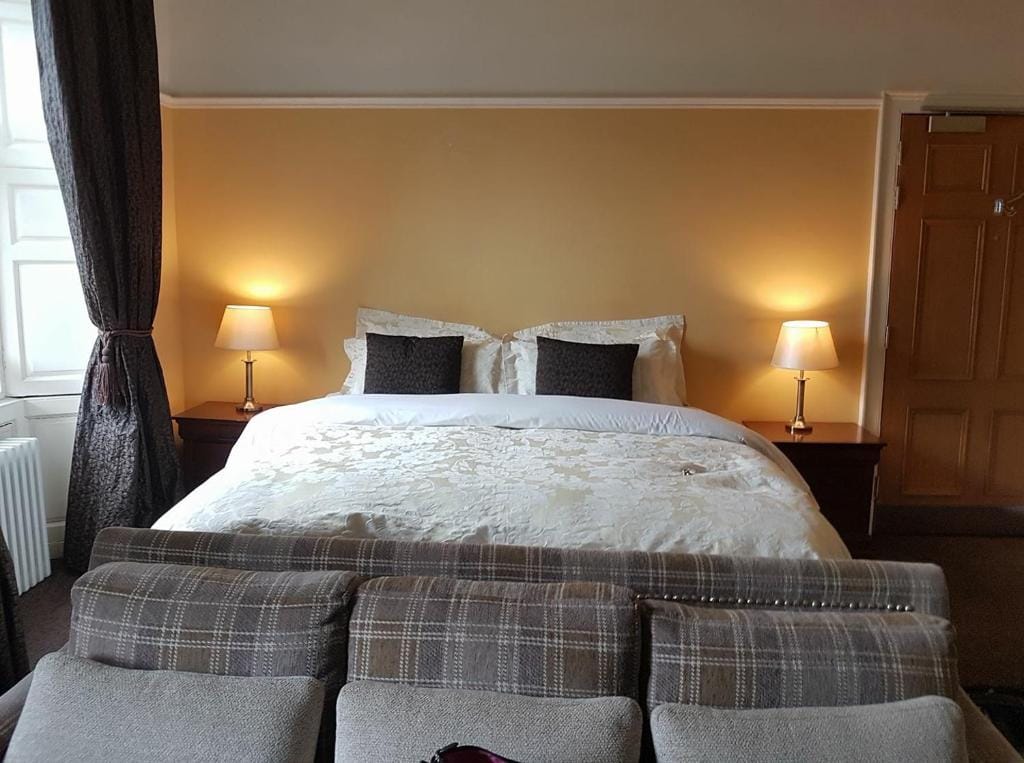 A cozy bedroom with a large bed, white bedding, and dark accent pillows. There are two bedside tables with lamps, a plaid sofa at the foot of the bed, brown carpet, and tan walls with a wooden door on the right.