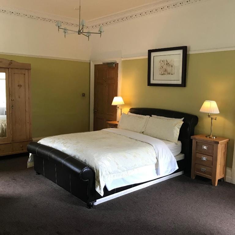 A neatly made bed with white linens sits between two wooden nightstands, each with a lamp. There’s a wardrobe on the left, a wooden door, greenish walls, and a framed picture above the bed in a cozy bedroom.