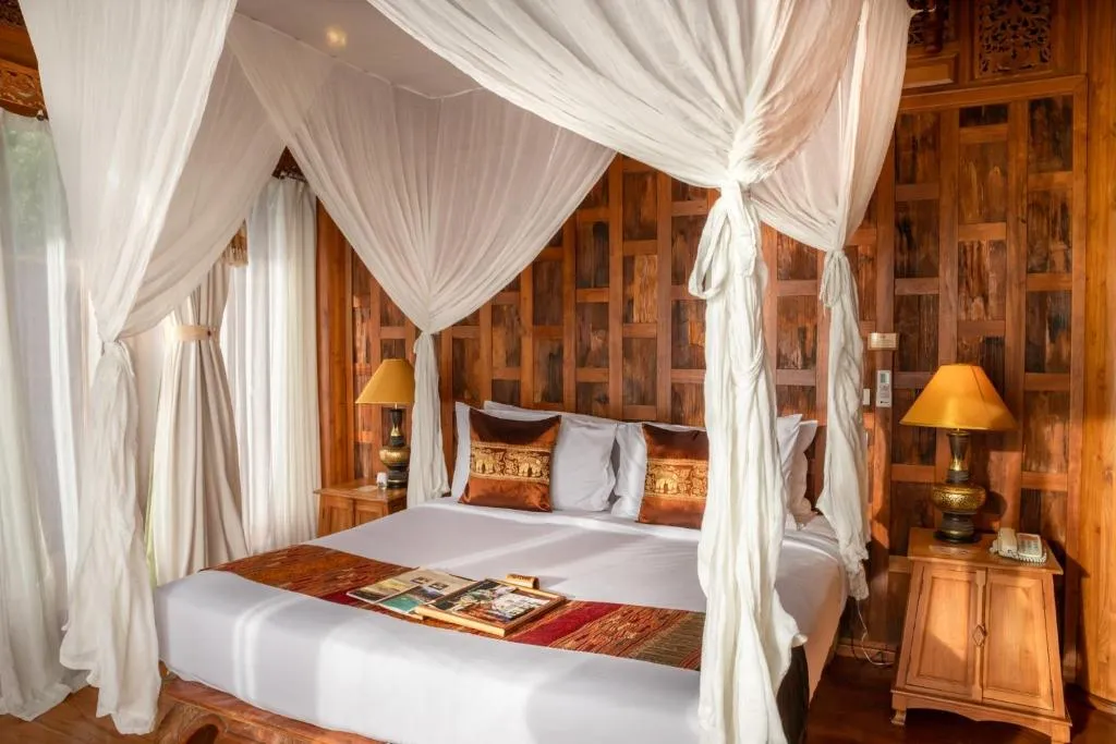 A cozy bedroom with a wooden canopy bed, sheer white drapes, decorative cushions, and bedside tables with lamps, set against a warm, wood-paneled wall. Magazines are placed on the neatly made bed.