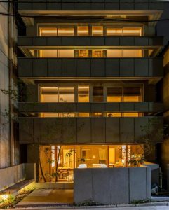 A modern multi-story building at night with large windows glowing with warm yellow light, revealing minimalistic interior spaces and some greenery near the entrance.