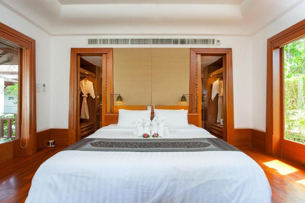 A bright, modern bedroom with a large neatly made bed, white linens, towel art, two open closets with robes hanging, wooden floors, and sunlit windows on both sides.