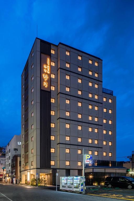 A tall, modern hotel building with many lit windows stands on a city street at dusk. The hotel has illuminated signage in Japanese and a parking area with cars in front. The sky is a deep blue.