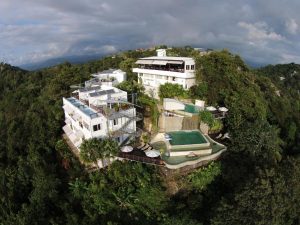 Aerial view of a large, modern white villa complex with a swimming pool, surrounded by dense green forest on a hillside under a cloudy sky.