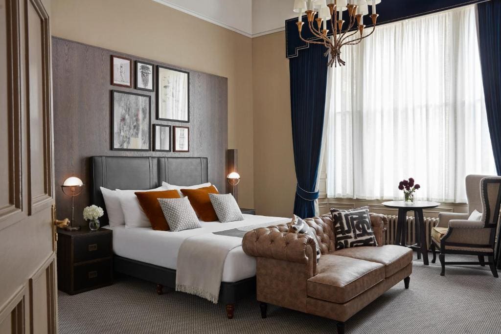 Stylish hotel room with a double bed, brown velvet bench, decorative pillows, armchair, round table with chairs, large window with dark blue curtains, wall art, and chandelier. Warm, elegant, and inviting atmosphere.