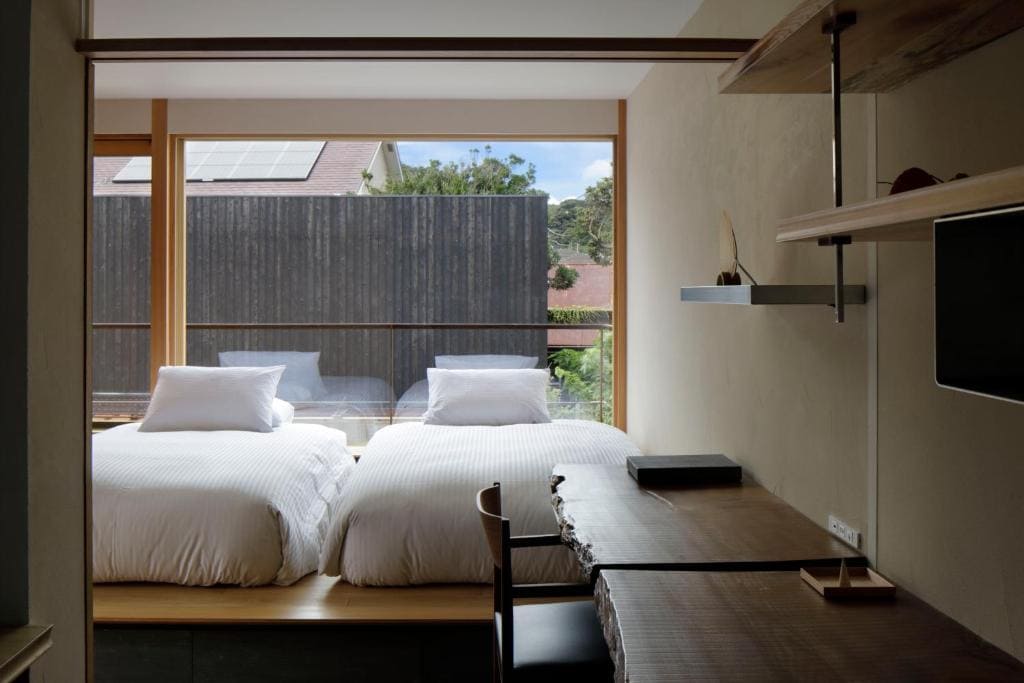 A minimalist bedroom features two twin beds with white bedding beside a large window, a wooden desk with shelves, and a view of rooftops and greenery outside.