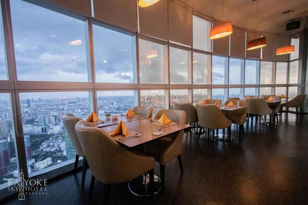 A modern restaurant with beige chairs and neatly set tables overlooks a city skyline through large floor-to-ceiling windows at sunset. Warm pendant lights hang from the ceiling, creating an inviting atmosphere.