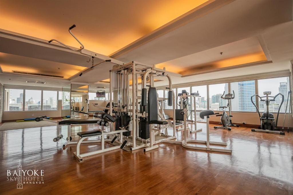 A modern hotel gym with wooden floors, large windows showing a city skyline, and various exercise machines including a weight station and cardio equipment. Warm ceiling lights create a welcoming atmosphere.