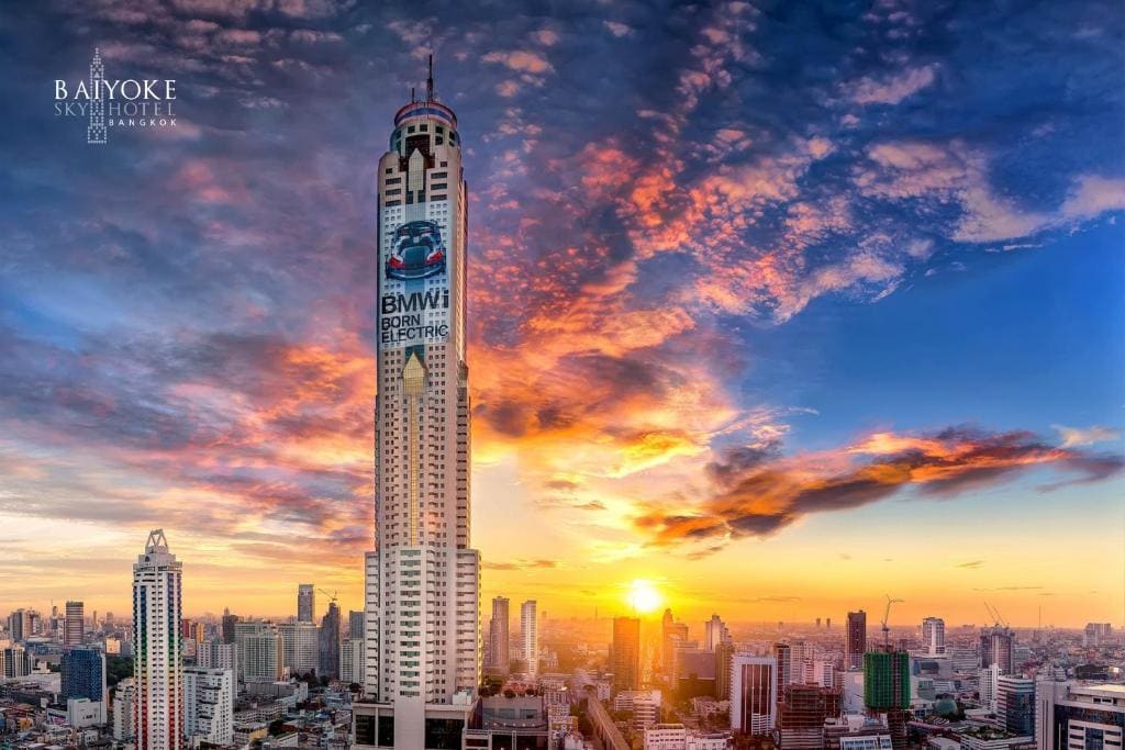 Tall Baiyoke Sky Hotel in Bangkok stands prominently among city buildings at sunset, with a colorful sky and dramatic clouds in the background. A BMW advertisement is visible on the tower.