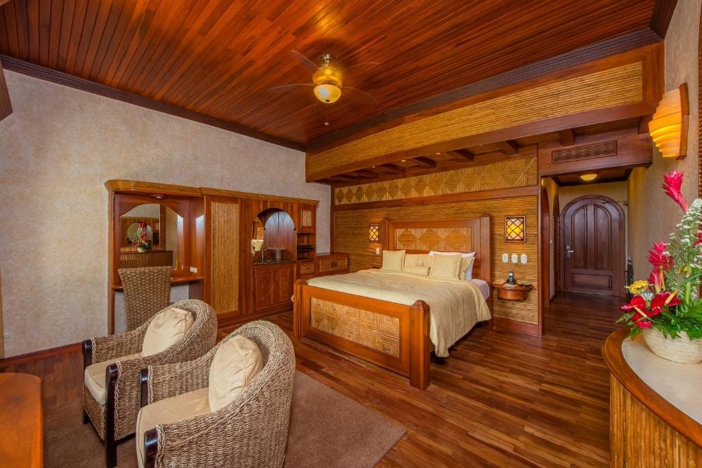 A cozy hotel room with a wooden ceiling and floor, a large bed with woven headboard, two wicker chairs, a small table, a wardrobe, and tropical flower arrangements. Warm lighting creates a welcoming atmosphere.